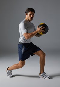 Athlet i tætsiddende grå t-shirt med korte ærmer og marineblå shorts, holder en sort og gul medicinbold. Grå baggrund, atletiske sko synlige.