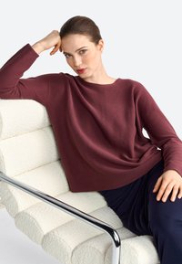Pull en tricot bordeaux à manches longues et à la silhouette décontractée, associé à un pantalon bleu marine, assis sur une chaise blanche texturée avec des bras en chrome.