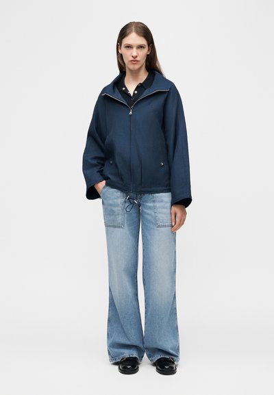 Γυναίκα που φοράει ναυτικό μπλε τζάκετ με φερμουάρ, ανοιχτό μπλε τζιν παντελόνι με φαρδιά μπατζάκια και μεγάλες μπροστινές τσέπες, και μαύρα παπούτσια, στέκεται με τα χέρια στις τσέπες.