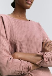 Světle růžový textured svetr s kulatým výstřihem a dlouhými rukávy, které mají nabírané manžety zavázané mašličkami, zdůrazňující útulný design.