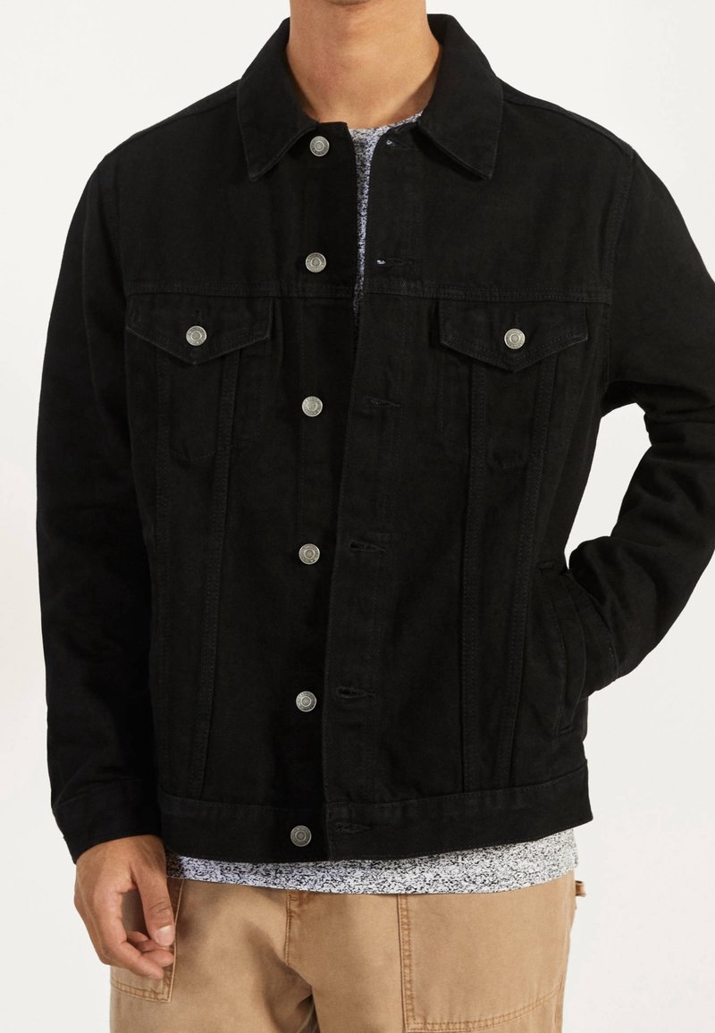 Svart denimjacka med silverfärgade tryckknappar, två bröstfickor och sidfickor. Har en ren design och texturerad tyg.