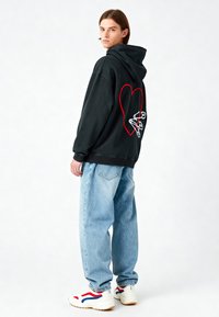 Felpa nera con grafica a cuore rosso e spille di sicurezza bianche sul retro, abbinata a jeans baggy azzurri chiari e sneaker bianche con dettagli rossi.