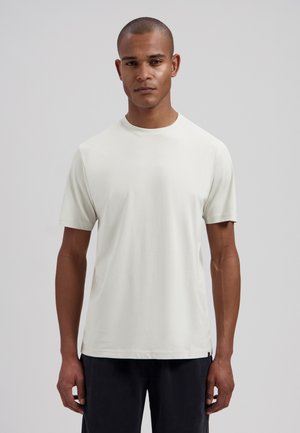 Man with short hair wearing plain white short-sleeve T-shirt and dark pants, standing against a plain light gray background.