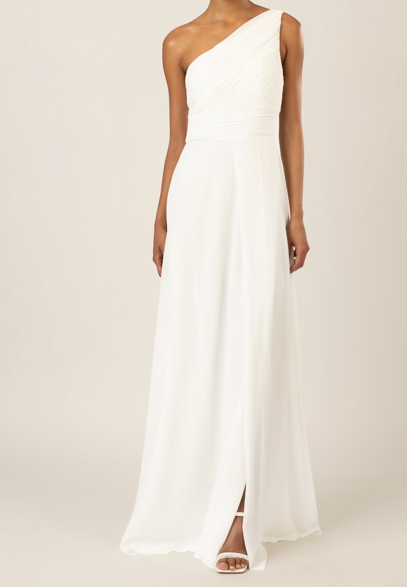 Femme portant une robe blanche longue au sol, une épaule dénudée, avec une fente montrant une sandale blanche à un pied.