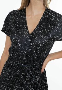 Svart omlottklänning med korta ärmar, prydd med vita prickar. Mjuk, texturerad tyg. Knytband vid midjan.