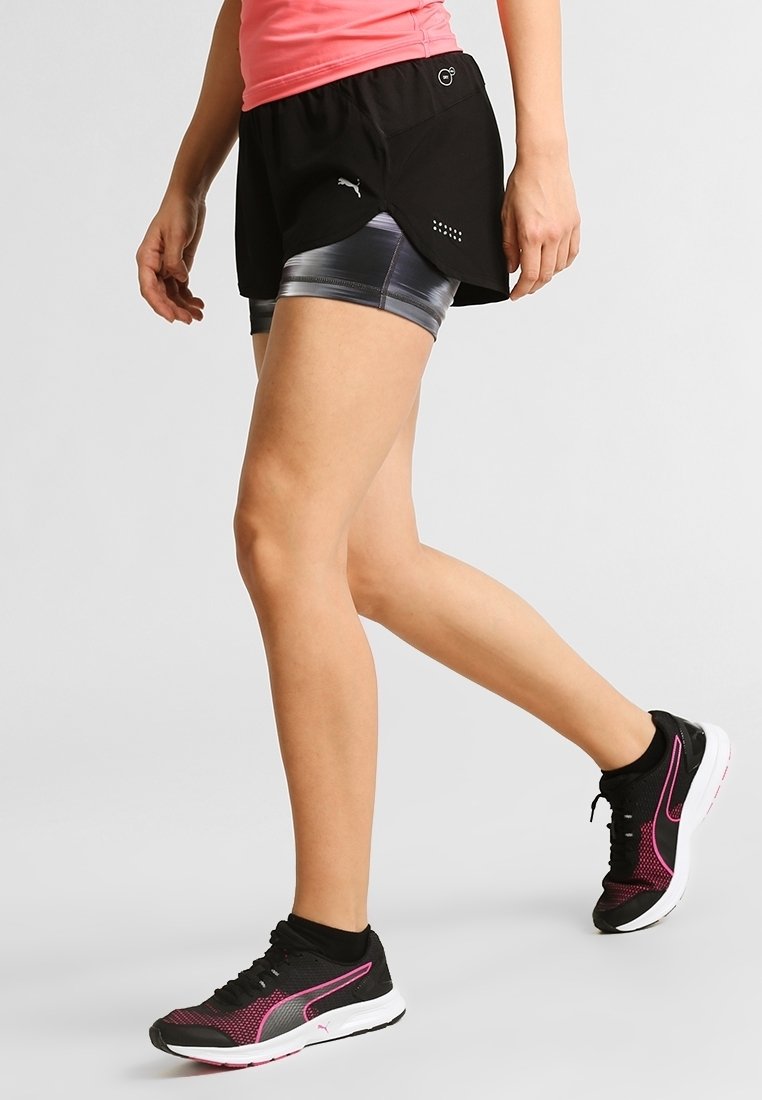 Pantalones cortos deportivos negros con un diseño en capas sobre pantalones cortos interiores grises brillantes. Usados con zapatillas de correr negras y rosas.