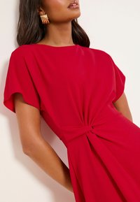 Červené šaty s voľným strihom, krátkymi rukávmi a uzlovým detailom v páse. Hladká látka s jemným záhybom. Zlaté kruhové náušnice.