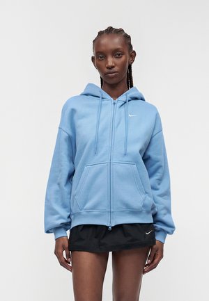 Žena v svetlomodrej mikine na zips a čiernej sukni s logami Nike stojí pred jednofarebným bielym pozadím.