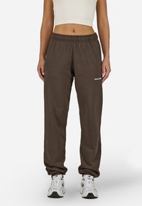 Brūnas sporta bikses no mīksta auduma ar elastīgu jostu, ar baltu logotipu kreisajā augšstilbā un sašaurinātiem piedurkņu galiem.