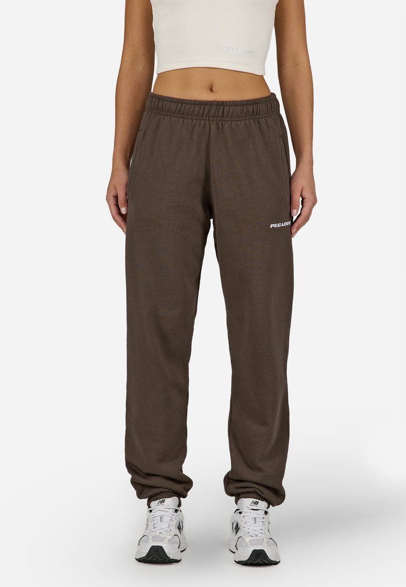 Brūnas sporta bikses no mīksta auduma ar elastīgu jostu, ar baltu logotipu kreisajā augšstilbā un sašaurinātiem piedurkņu galiem.