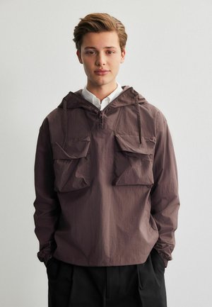 Chaqueta con capucha marrón hecha de una tela ligera, presenta dos bolsillos frontales, cordones ajustables y puños elásticos.