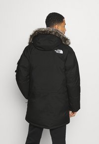 Zwarte parka-jack met een capuchon met bontafwerking, voorzien van een logopatch op de schouder, een langere snit en met ritszakken op de mouwen.