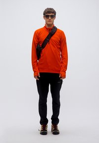 Πορτοκαλί πουλόβερ με μακριά μανίκια, μαύρο παντελόνι και ορειβατικά μποτάκια. Αξεσουάρ: μαύρη τσάντα χιαστί. Το μοντέλο φορά γυαλιά ηλίου, στέκεται μπροστά σε απλό φόντο.