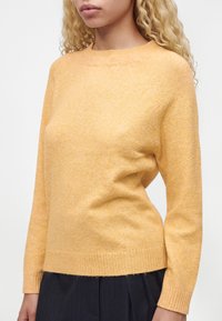 Woman wearing a soft yellow crew neck sweater with long sleeves and ribbed cuffs paired with dark pinstriped pants.