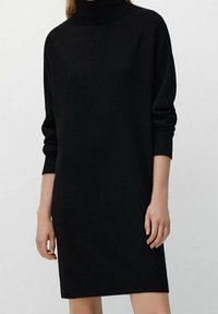 Femme portant une robe-pull noire à col roulé à manches longues, debout avec les bras détendus le long du corps sur un fond uni.
