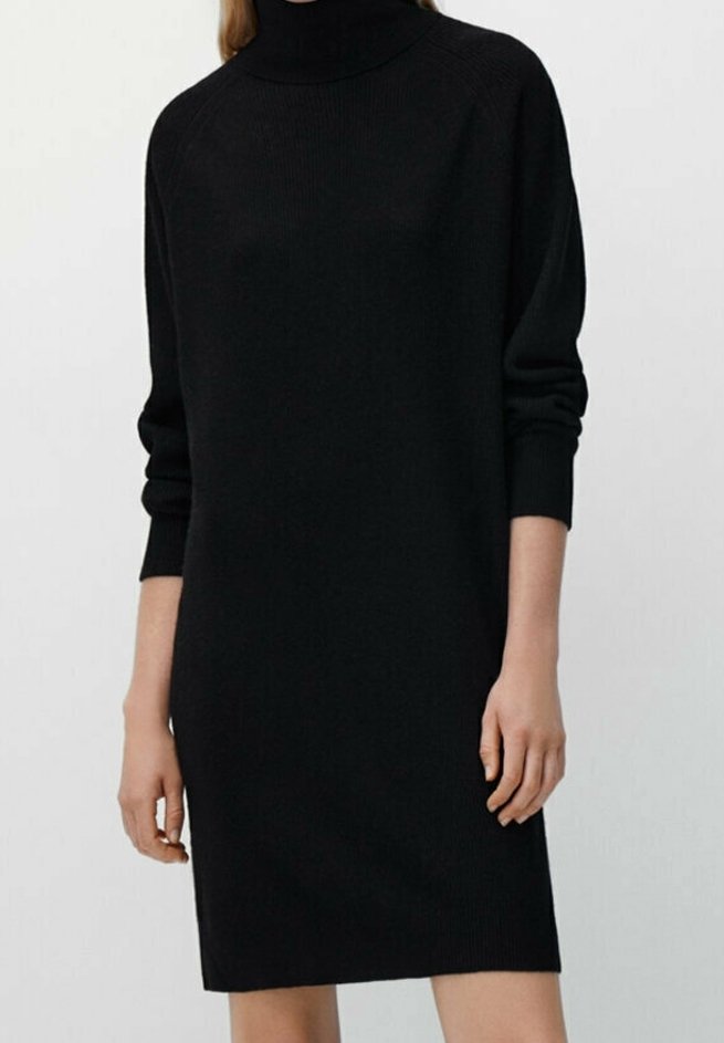 Femme portant une robe-pull noire à col roulé à manches longues, debout avec les bras détendus le long du corps sur un fond uni.