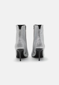 Botas de tobillo plateadas con un acabado brillante, tacones negros y cremalleras en el lateral. Textura suave y diseño de punta afilada.