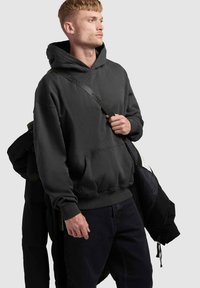 Svart hoodie i mjukt tyg med en framficka, ribbade ärmar och en justerbar snörning i huvan. Bärs med mörka jeans och en jacka.