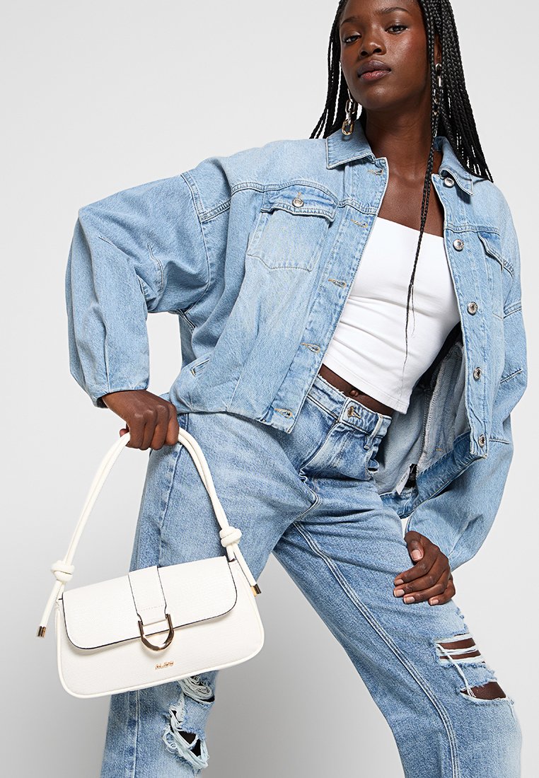 Jeune femme aux longues tresses porte une veste en jean bleu clair et un jean déchiré, tenant un sac à main en cuir blanc avec des sangles nouées.