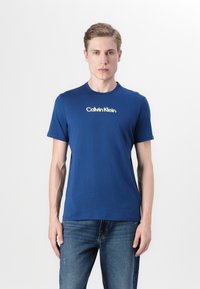 Μπλε βαμβακερό μπλουζάκι με κοντά μανίκια και στρογγυλή λαιμόκοψη, που διαθέτει λευκό λογότυπο σε αντίθεση στο κέντρο. Φορεμένο με μπλε τζιν.