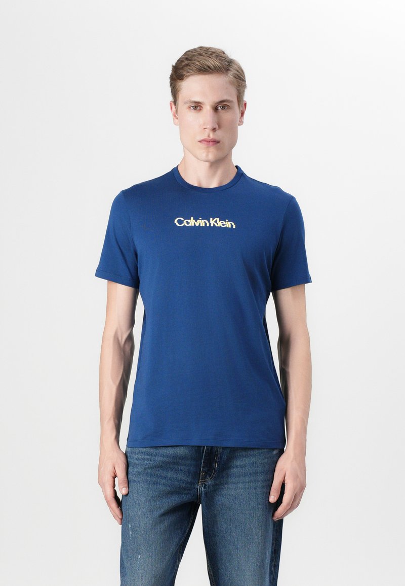 Μπλε βαμβακερό μπλουζάκι με κοντά μανίκια και στρογγυλή λαιμόκοψη, που διαθέτει λευκό λογότυπο σε αντίθεση στο κέντρο. Φορεμένο με μπλε τζιν.