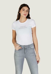 Jonge vrouw met lang donker haar, gekleed in een wit Armani Exchange T-shirt en lichtgrijze spijkerbroek, staand tegen een effen achtergrond.