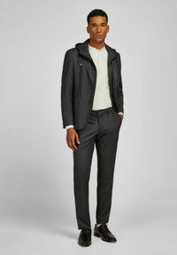 Donkergrijs op maat gemaakt pak met een capuchonjack van een gestructureerde stof. Slim-fit broek en een lichtgekleurde henley-shirt eronder. Zwarte nette schoenen.