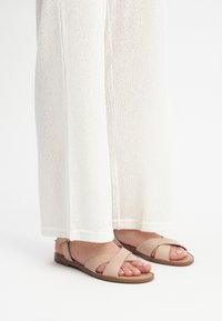 Beżowe płaskie sandały z krzyżującymi się paskami. Wykonane z miękkiej skóry, o minimalistycznym stylu. Noszone z białymi, luźnymi dzianinowymi spodniami.