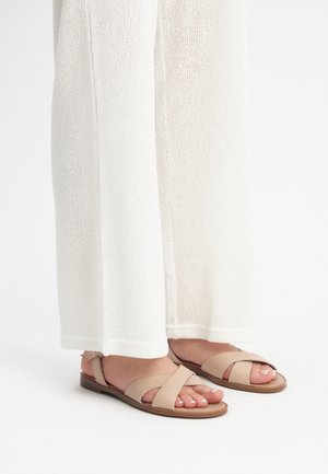 Beżowe płaskie sandały z krzyżującymi się paskami. Wykonane z miękkiej skóry, o minimalistycznym stylu. Noszone z białymi, luźnymi dzianinowymi spodniami.