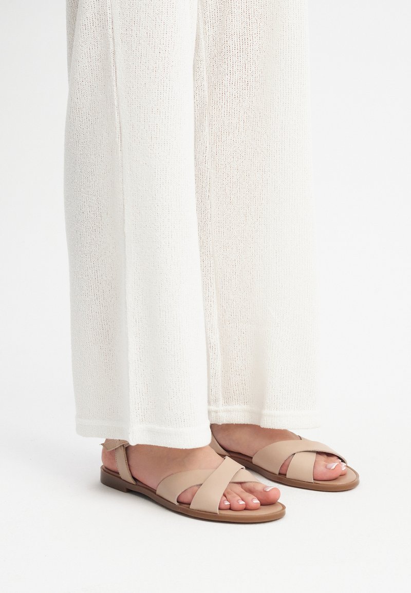 Beżowe płaskie sandały z krzyżującymi się paskami. Wykonane z miękkiej skóry, o minimalistycznym stylu. Noszone z białymi, luźnymi dzianinowymi spodniami.