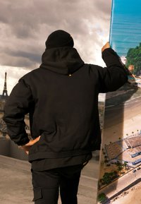 Giacca nera con cappuccio, dalla vestibilità rilassata e orlo a costine, abbinata a pantaloni scuri coordinati. La figura tiene un poster colorato.