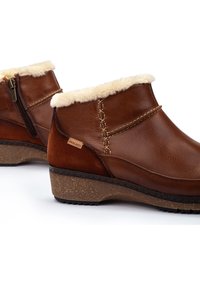 Botas de tobillo de cuero marrón con un cuello de piel sintética, cremallera lateral, costuras decorativas y una suela de cuña texturizada. Logo de Pikolinos visible.