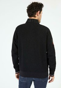 Sweatshirt noir avec un col montant, des poignets côtelés et un logo à l'arrière. Présente des rayures contrastantes à l'ourlet. Texture douce.