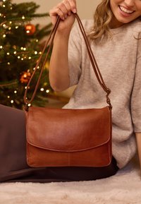 Smeđa kožna torba preko tijela s preklopom, podesivim remenom, glatkom teksturom i minimalističkim dizajnom, postavljena na udobnu, blagdansku pozadinu.