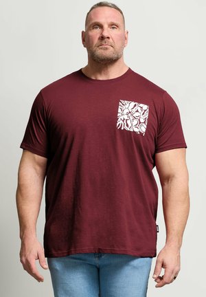 Muskelöser Mann trägt ein dunkelrotes T-Shirt mit einer weißen, floral gemusterten Tasche, steht mit entspannten Armen, heller Hintergrund.