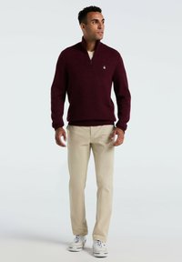 Burgunderfarbener Strickpullover mit Viertel-Reißverschluss-Kragen, beigen Hosen und weißen Sneakers. Der Pullover hat ein kleines Logo auf der Brust.