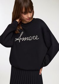 Czarna bluza z szerokimi rękawami i prążkowanym dekoltem, z białym haftem "Amore" na przodzie. Teksturowana tkanina, luźny krój.