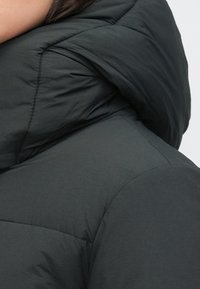 Sötét színű steppelt kabát magas gallérral, elegáns dizájnnal és visszafogott matt felülettel. Feltűnő varrási részletek a varratok mentén.