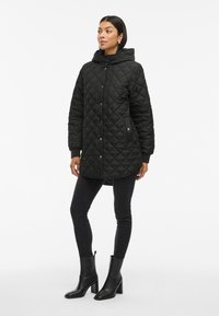 Parka acolchada negra con capucha, cierre de botones a presión, puños elásticos y bolsillos laterales, llevada con jeans ajustados negros y botines de tacón.