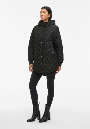 Zwarte gewatteerde parka met een capuchon, drukknoopsluiting, elastische boorden en zijzakken, gedragen met zwarte skinny jeans en hakschoenen.