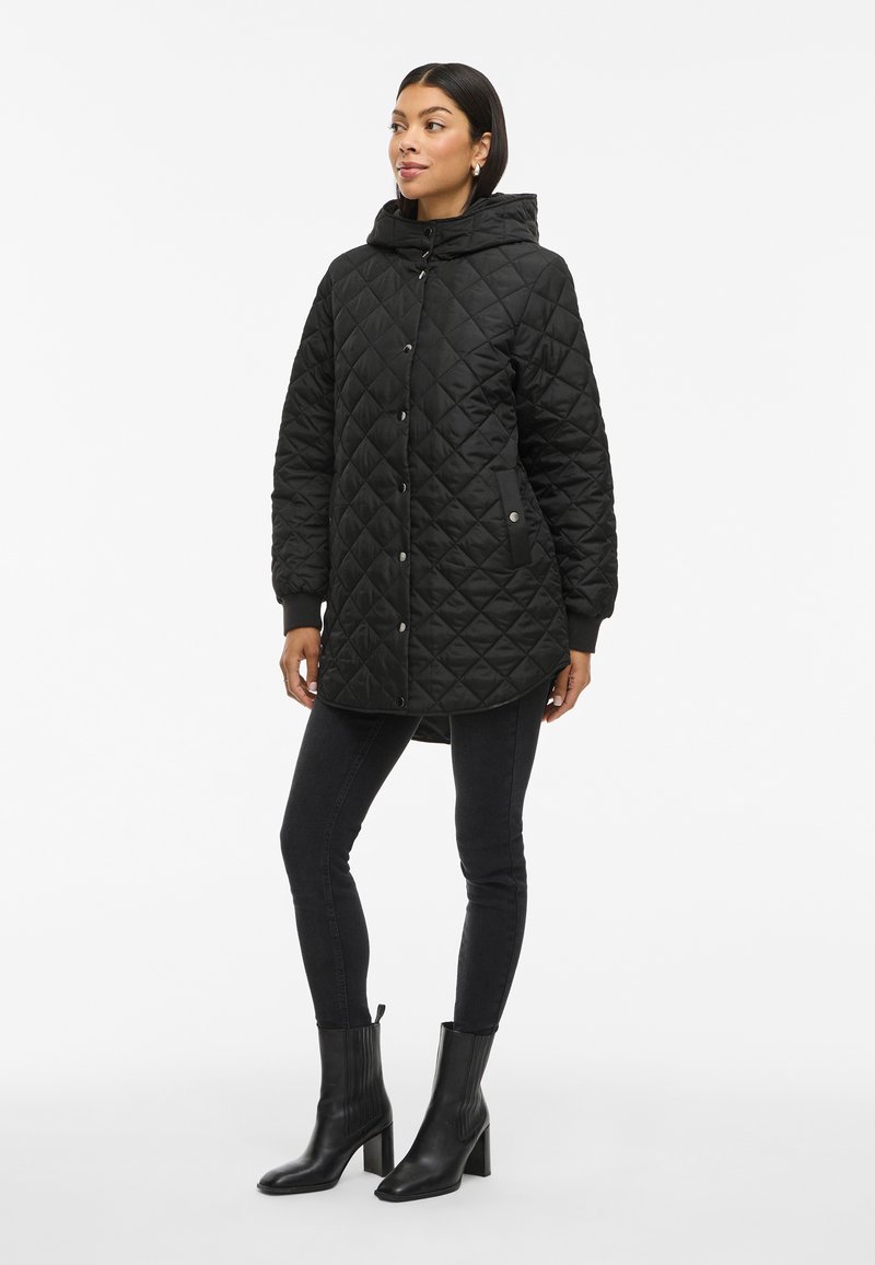 Parka acolchada negra con capucha, cierre de botones a presión, puños elásticos y bolsillos laterales, llevada con jeans ajustados negros y botines de tacón.