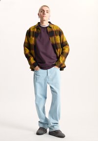 Camicia a quadri gialla e nera in flanella, felpa viola, jeans azzurri chiari, sneakers grigie. Outfit casual a strati con tasche e stampe.