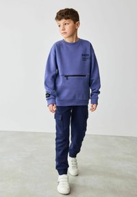 Lilla dressipluus, millel on ees kinnitustasku, logotekst ja tumesinised cargo-püksid; täiustatud valgete spordijalatsidega, millel on velcro rihmad.
