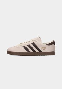 Beige suède Adidas sneaker met donkerbruine zool en drie donkerbruine strepen, veterluiting aan de voorkant en "Adidas Stadt" logo aan de hielkant.