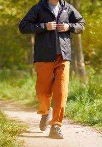 Giacca impermeabile nera, t-shirt grigia, pantaloni arancioni con cuciture sulle ginocchia e scarpe da trekking nere, che cammina su un sentiero circondato da verde.