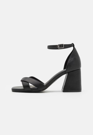 Μαύρες γυναικείες σандάλια με κλειστή πλάτη, λουράκι στον αστράγαλο και διασταυρωμένα μπροστινά λουριά. Κατασκευασμένα από λείο υλικό με χοντρό μπλοκ τακούνι.