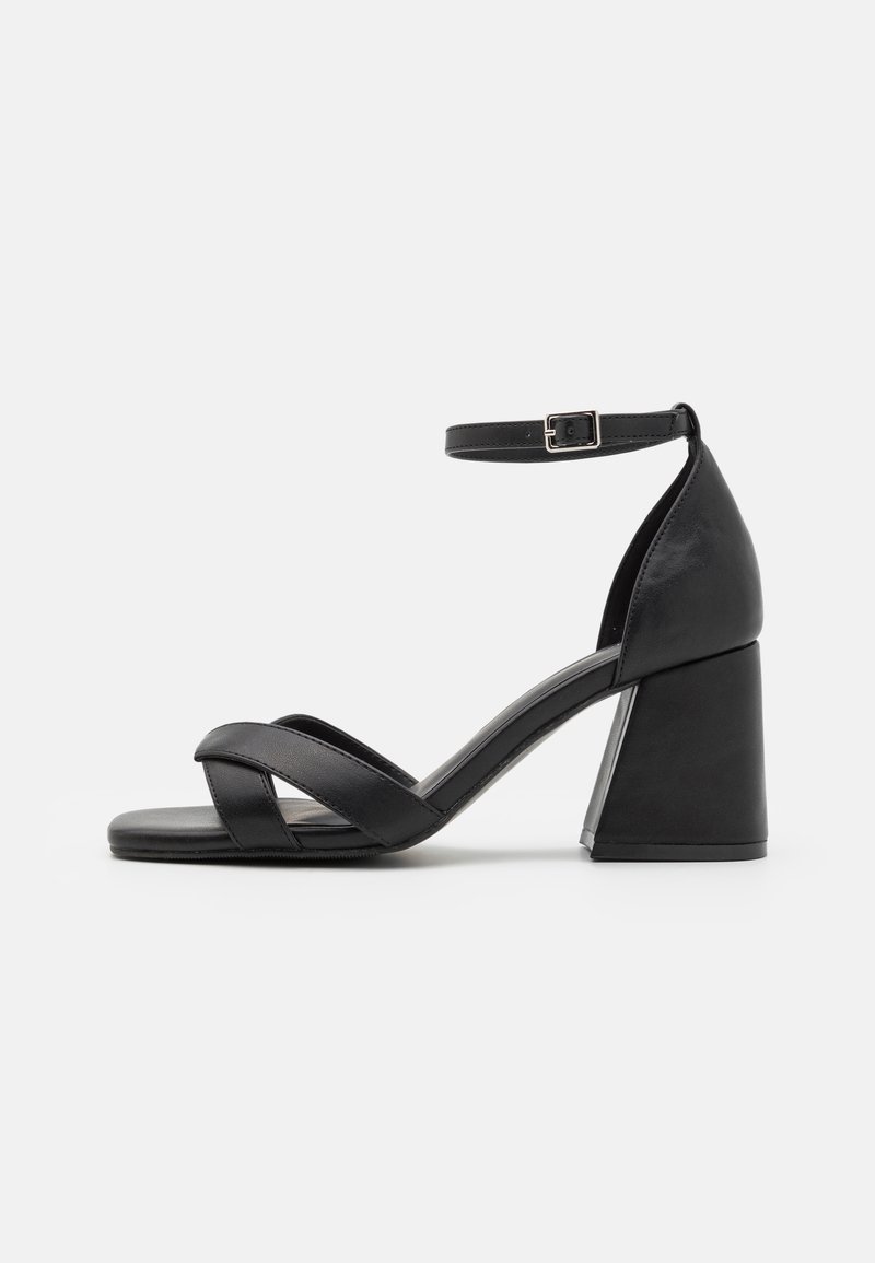 Svart hæl sandaler med lukket bak, ankelrem og krysset foran remmer. Laget av glatt materiale med en klumpete blokkhæl.