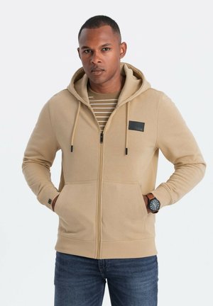 Sudadera con cremallera color beige, con capucha forrada, bolsillos frontales y un parche de logo negro. Llevada sobre una camiseta de rayas de color claro.