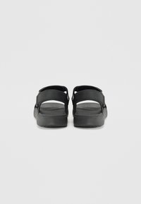 Svarte sandaler med to justerbare stropper, flate såler og teksturerte fotbed, sett fra baksiden. Mykt materiale med et minimalistisk design.