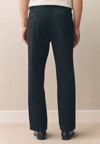 Pantalon noir à coupe droite, doté de poches arrière avec boutons, en tissu de coton avec une texture subtile. Porté avec des chaussures noires.